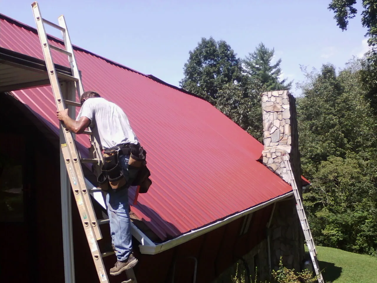 Roofer on a ladder beside a red metal roof and stone chimney — Soffit & Fascia Repair by Roofing Brothers in Crum Lynne